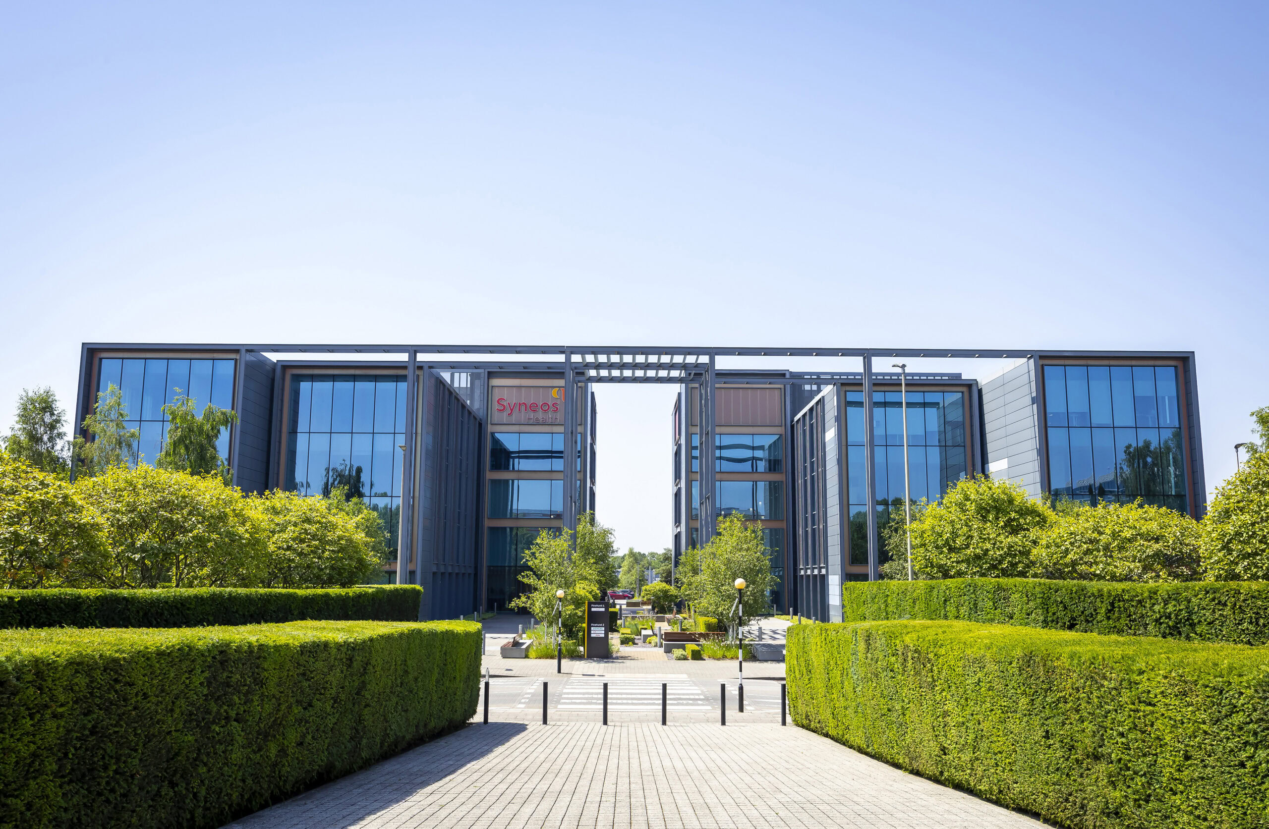 Modern office buildings with glass façades, surrounded by green hedges, trees, and a walkway.