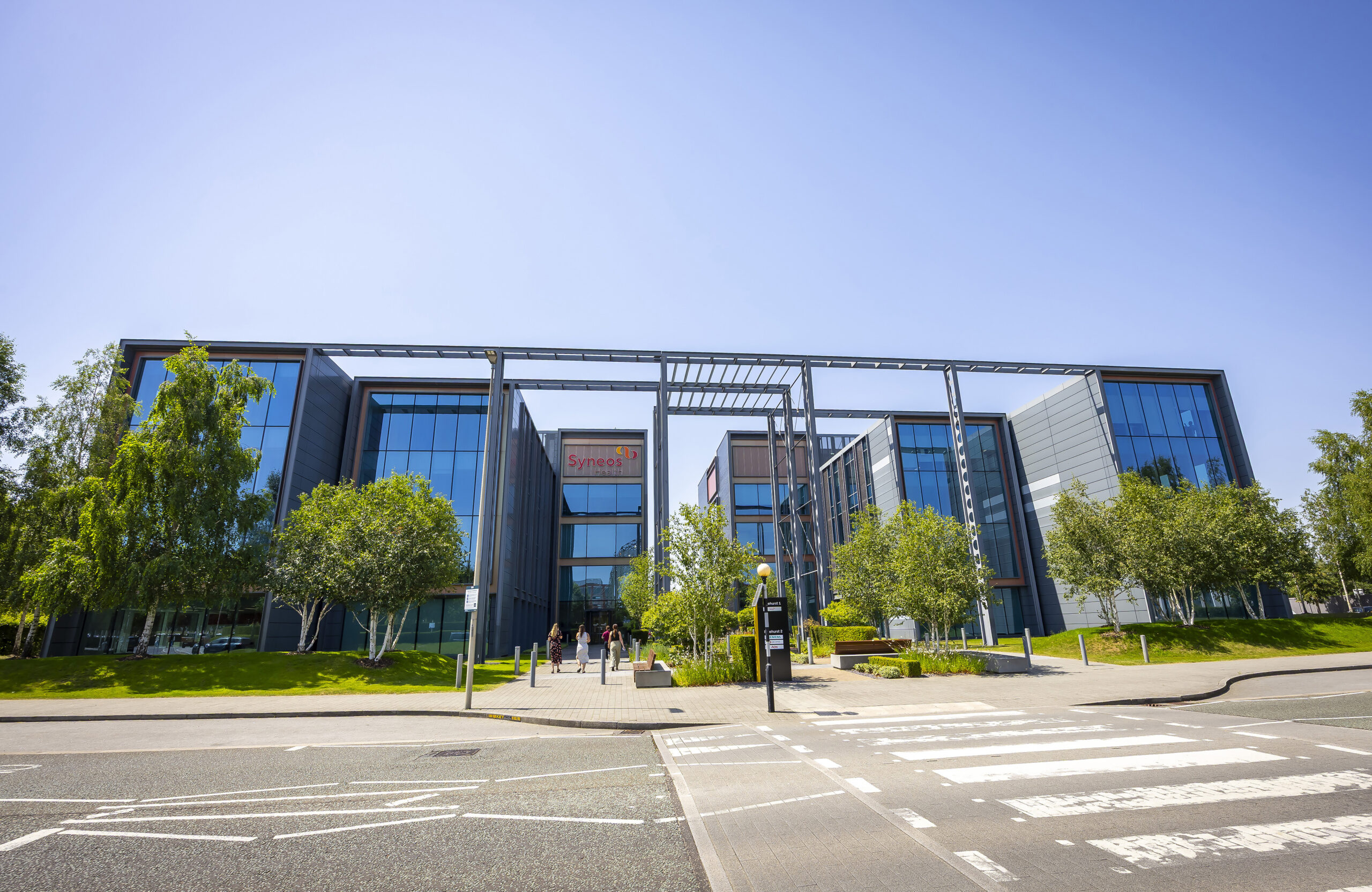 Modern glass office building with trees in front, viewed from across a street with a zebra crossing.