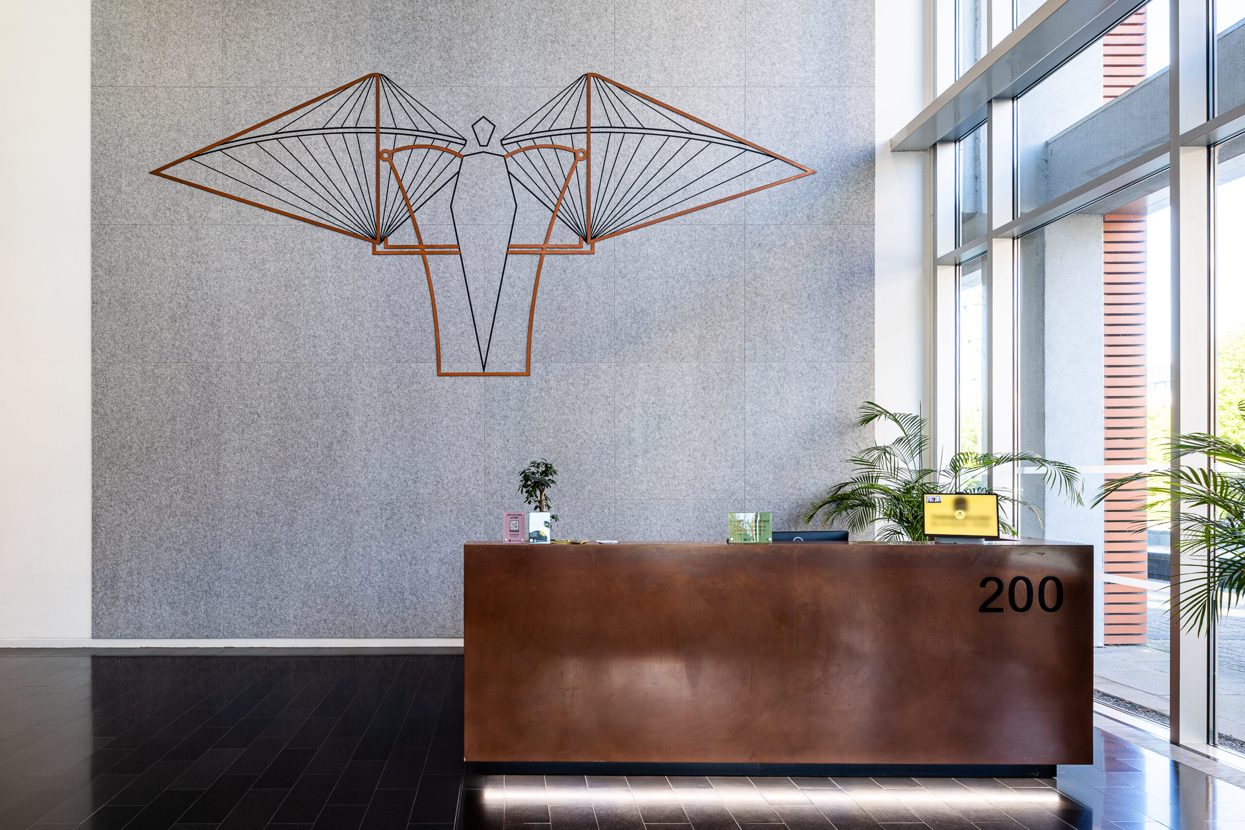 Modern office reception area with a metal desk, wall art, plants, and large windows.
