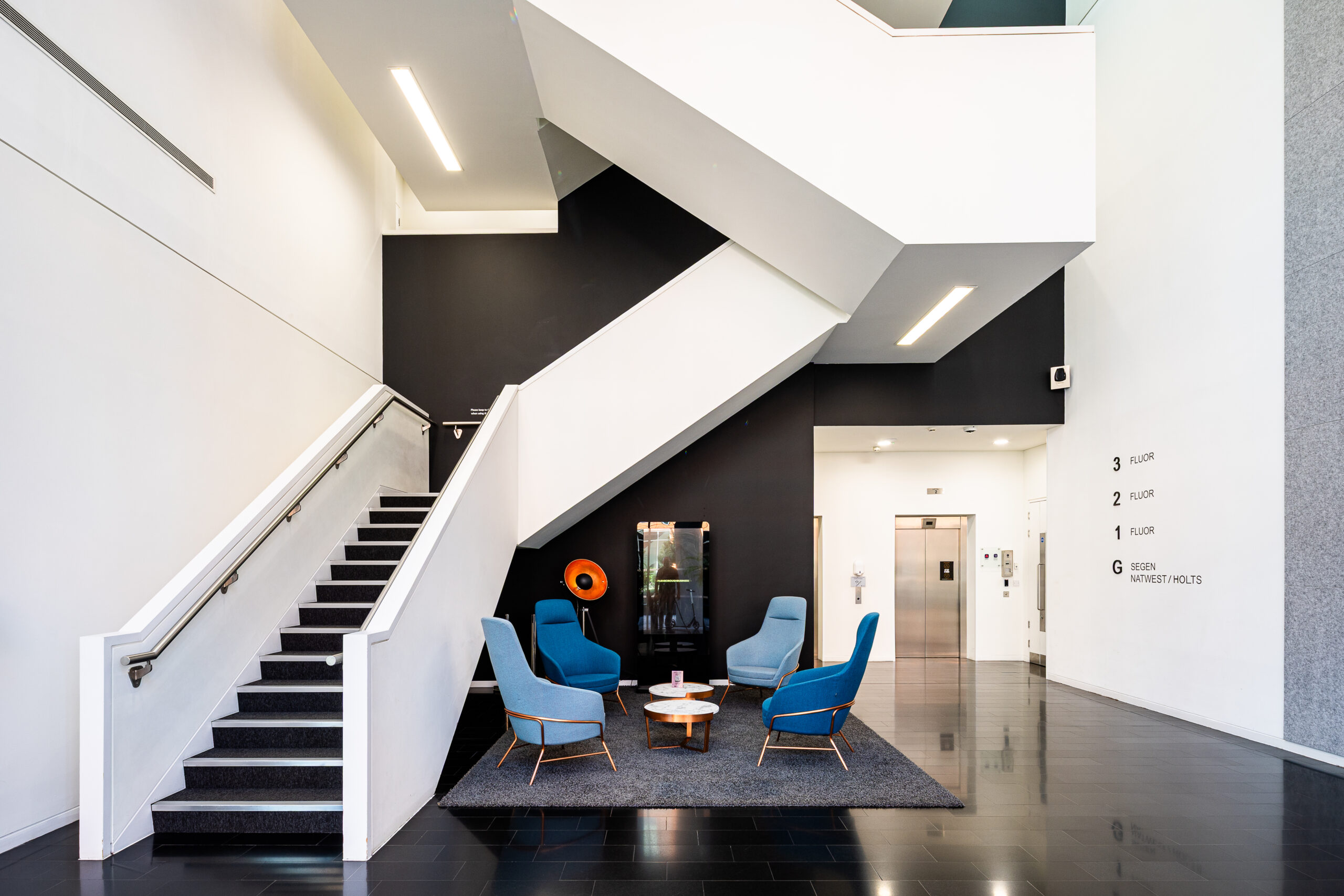 Modern lobby with blue chairs, a round table, and a staircase beside an elevator and floor directory.