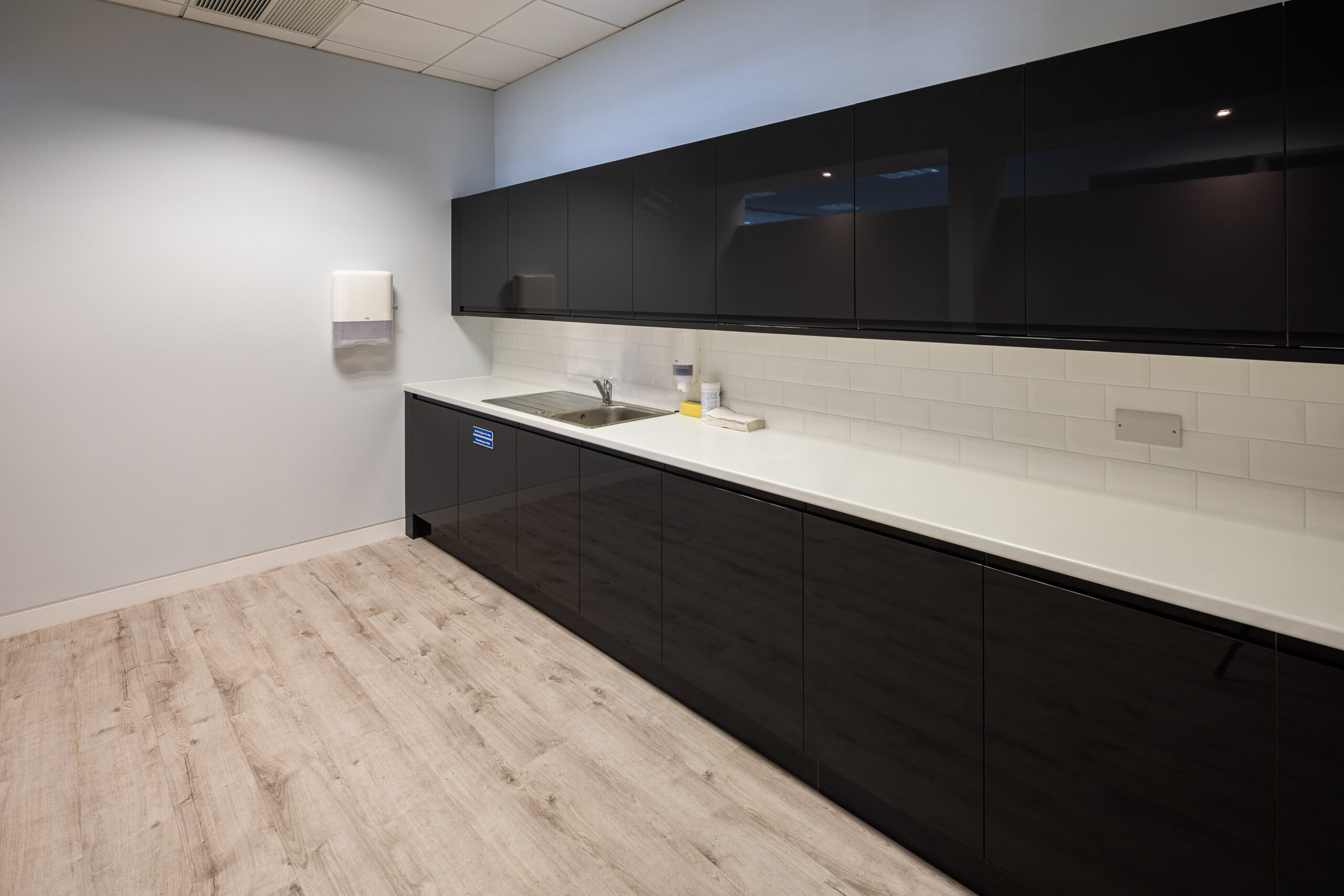Modern kitchen with black cabinets, white countertop, sink, and paper towel dispenser on the wall.