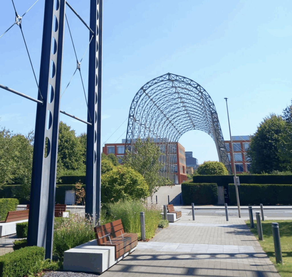 Tall metal framework and archway structure with benches and greenery on a sunny day.