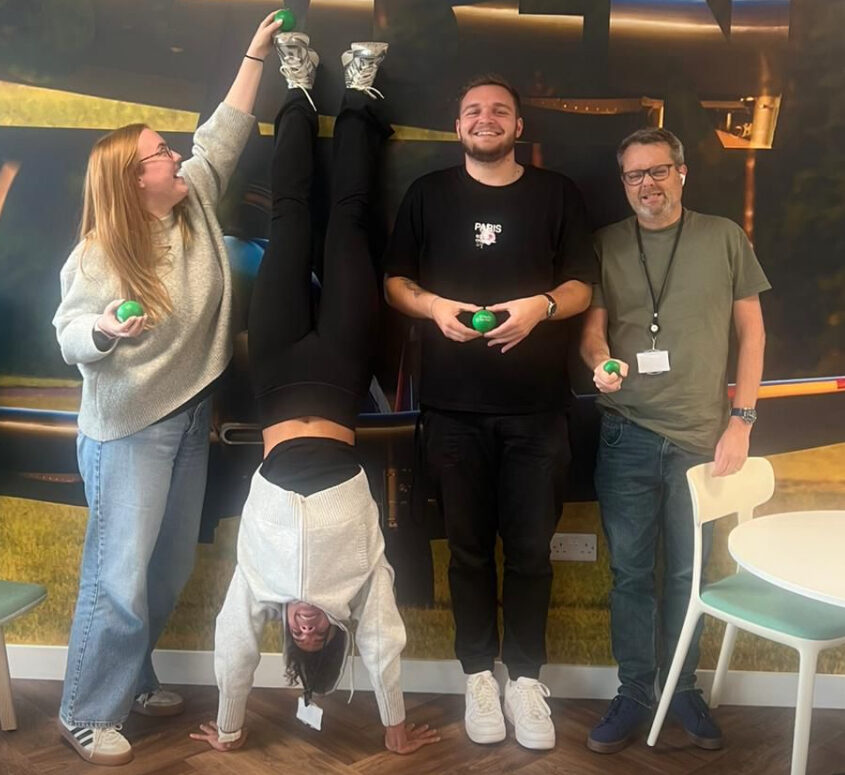 Three people stand while one person does a handstand indoors in front of a colorful wall mural.