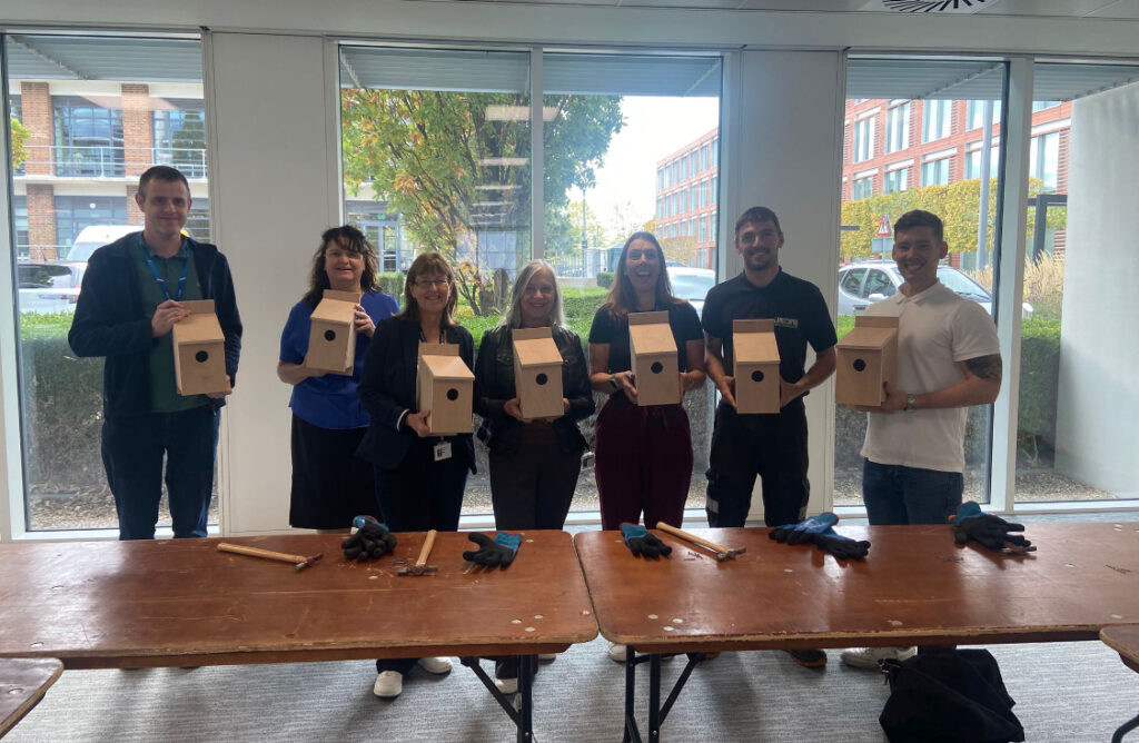 Seven people stand indoors holding wooden birdhouses, with tables and tools in the foreground.