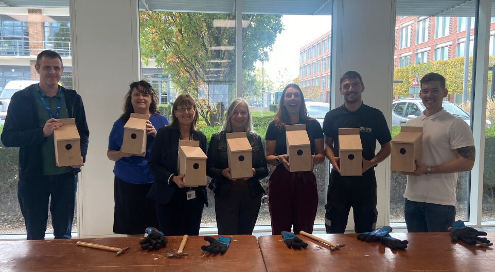 Seven people stand indoors holding wooden birdhouses, with tables and tools in the foreground.