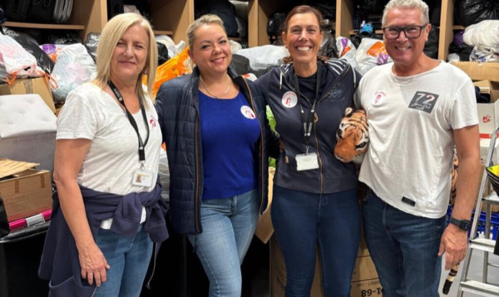 Four people stand together in a storeroom filled with boxes, bags, and assorted supplies.