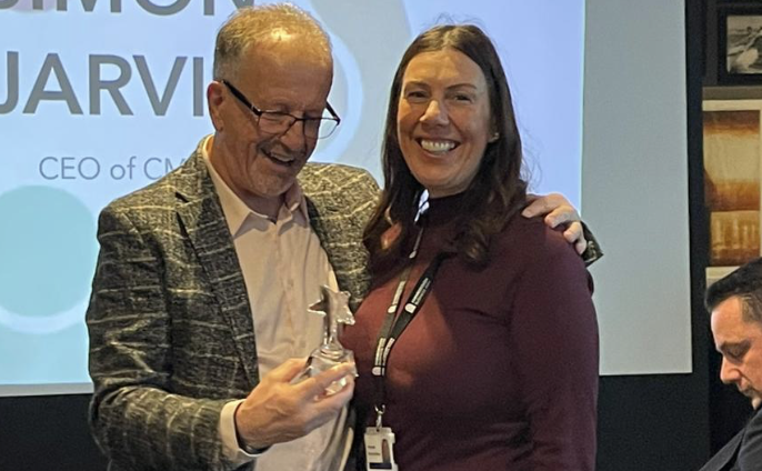 Two people stand together while one holds a glass award; others are seated in the background.