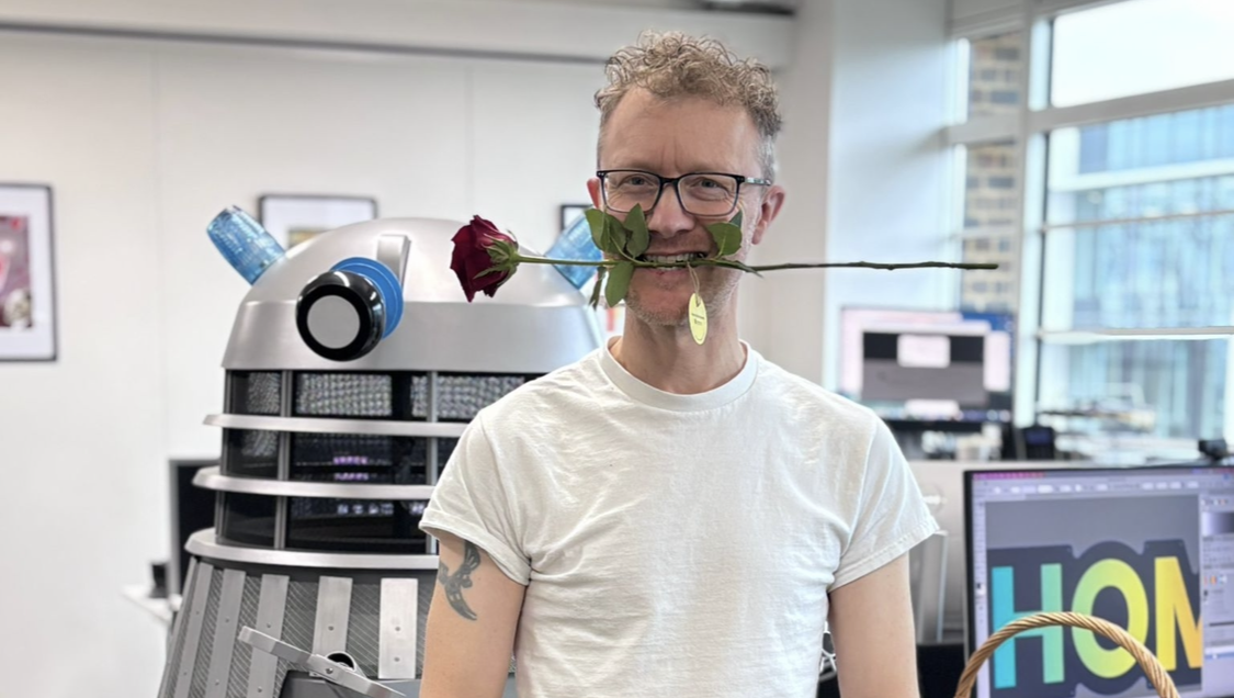A man with a rose in his mouth stands in an office beside a Dalek and a basket of roses.
