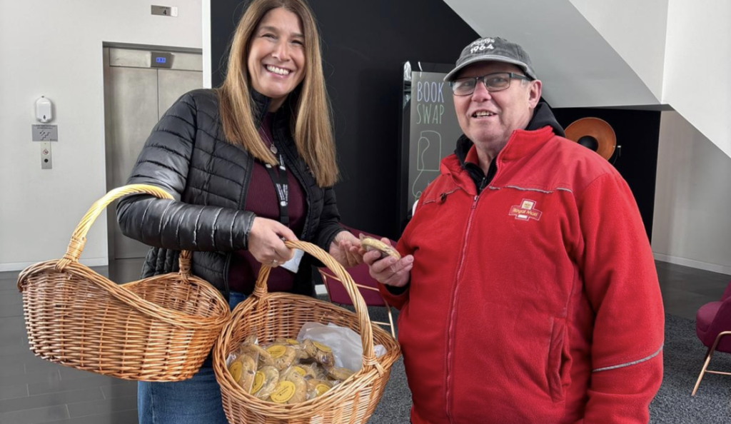 Two people stand indoors smiling, holding baskets filled with what appear to be packaged biscuits.