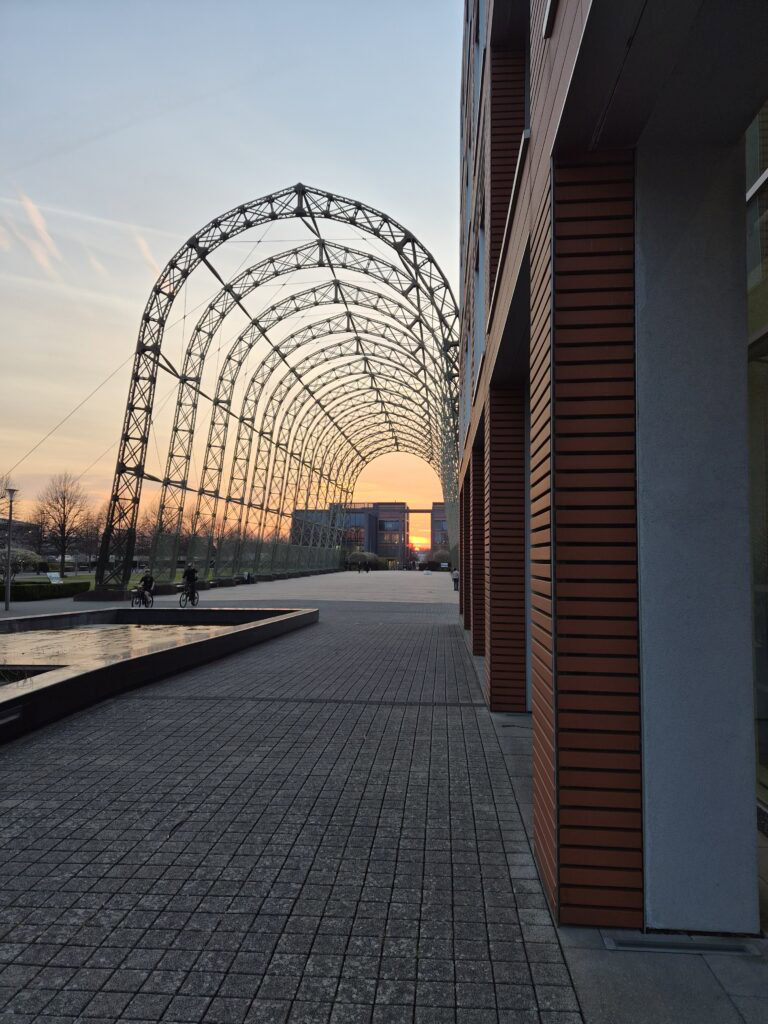 Arched metal structure and modern building at sunset with a paved walkway in the foreground.