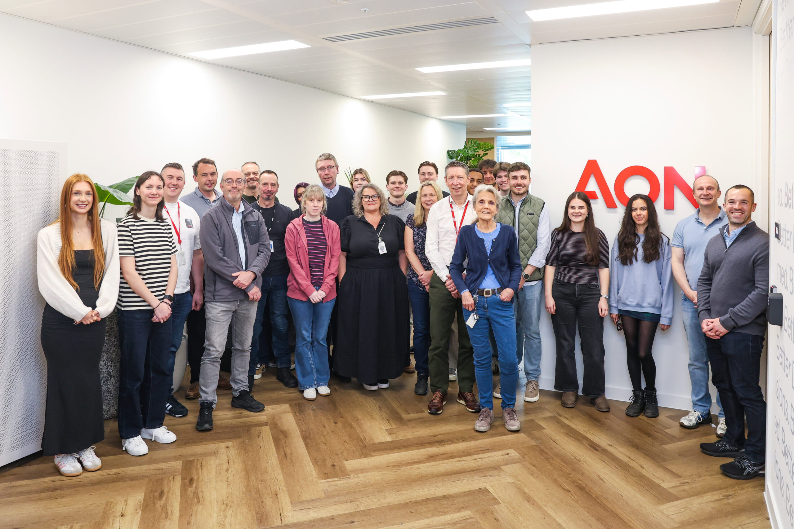 A group of people pose together in an office corridor next to an Aon sign on the wall.