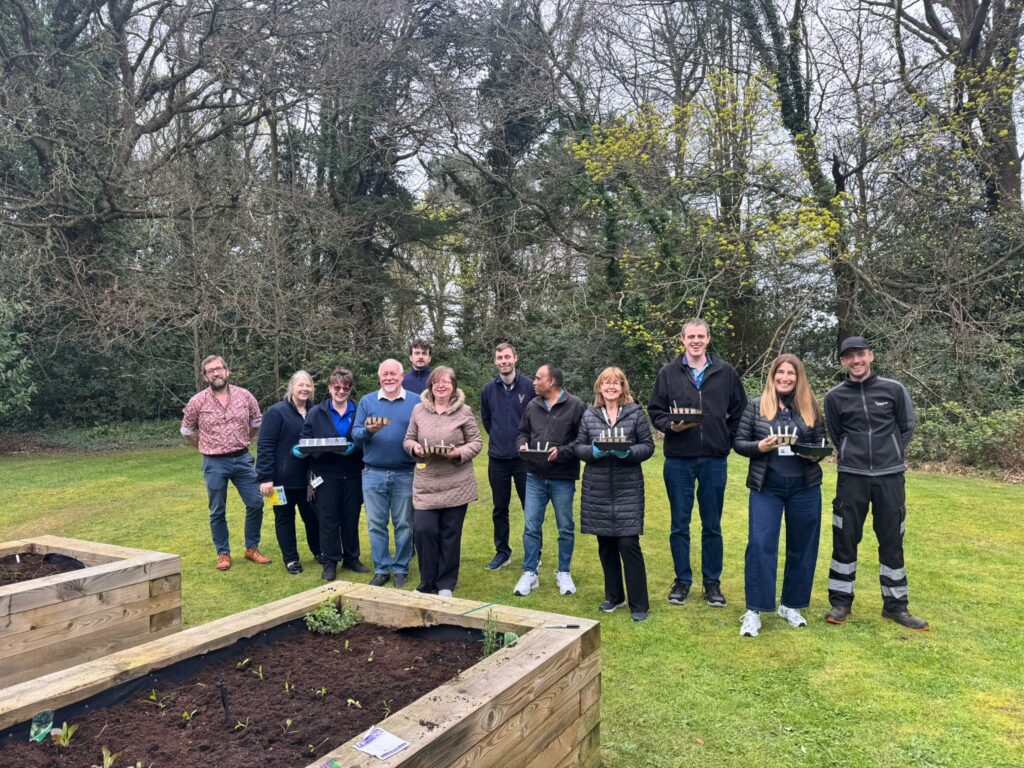 A group of people stand outdoors holding seed trays beside raised garden beds and trees.