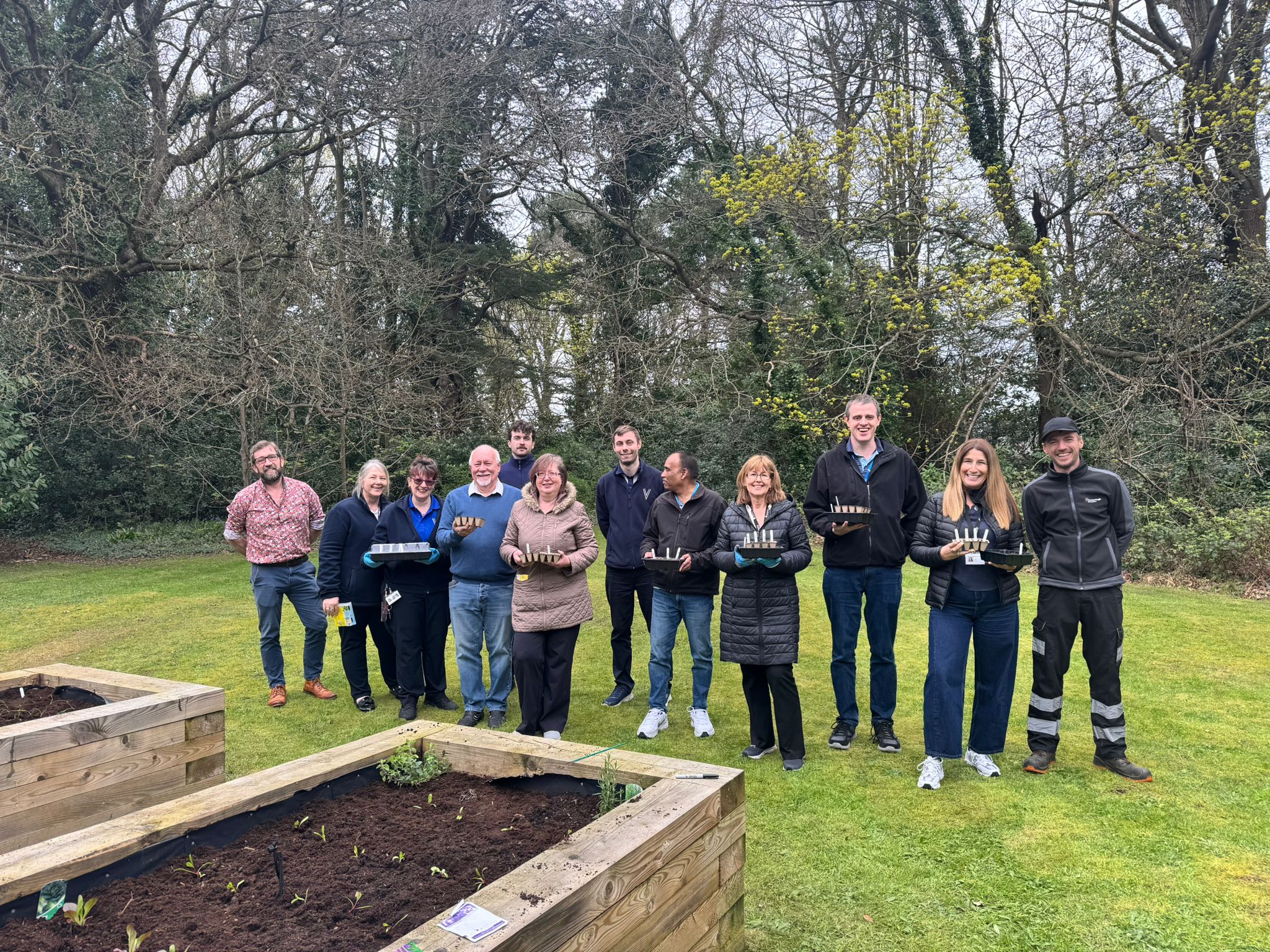 A group of people stand outdoors holding seed trays beside raised garden beds and trees.