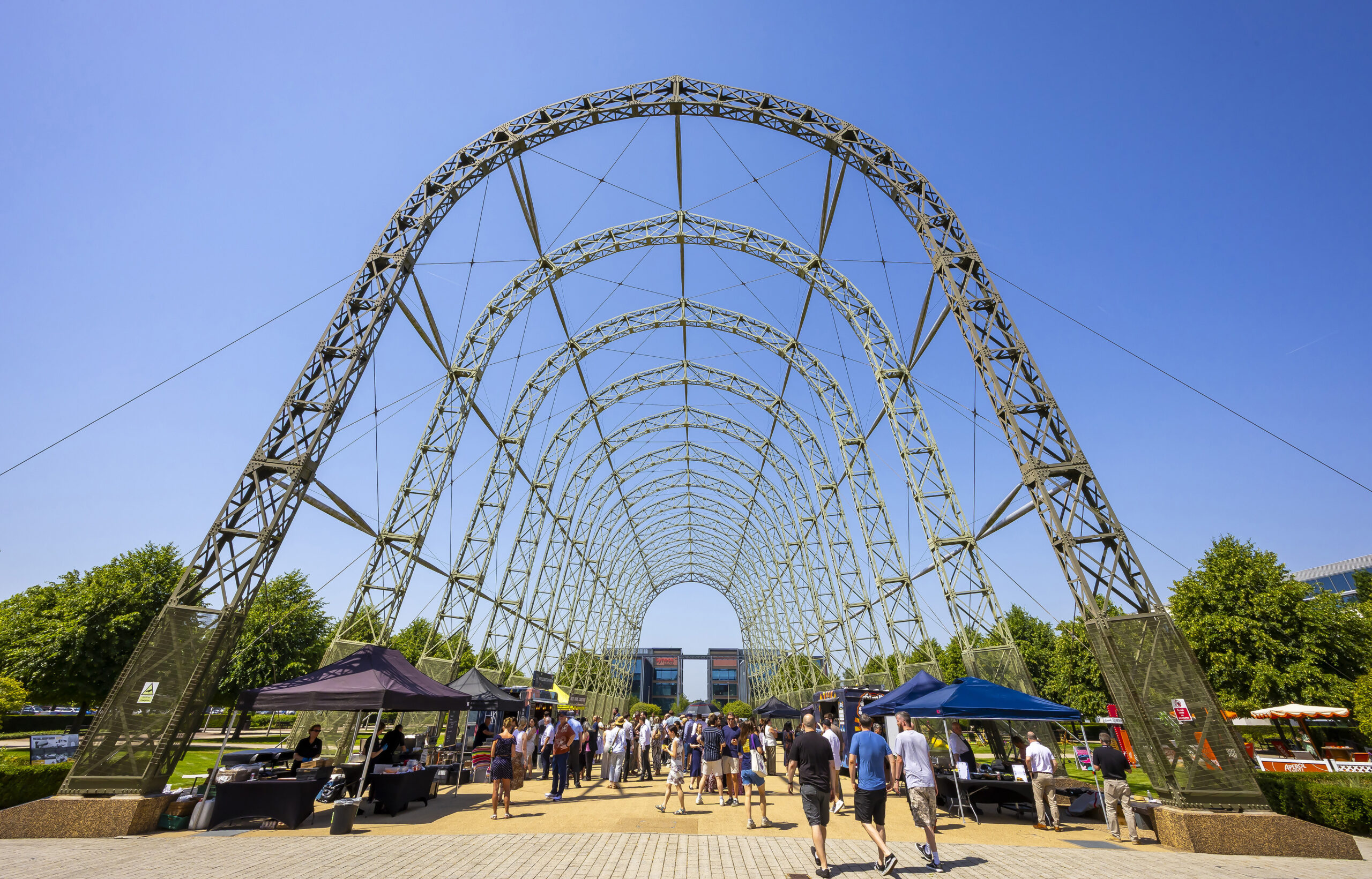 People walk under a large metal arch structure with vendor tents set up on both sides on a sunny day.