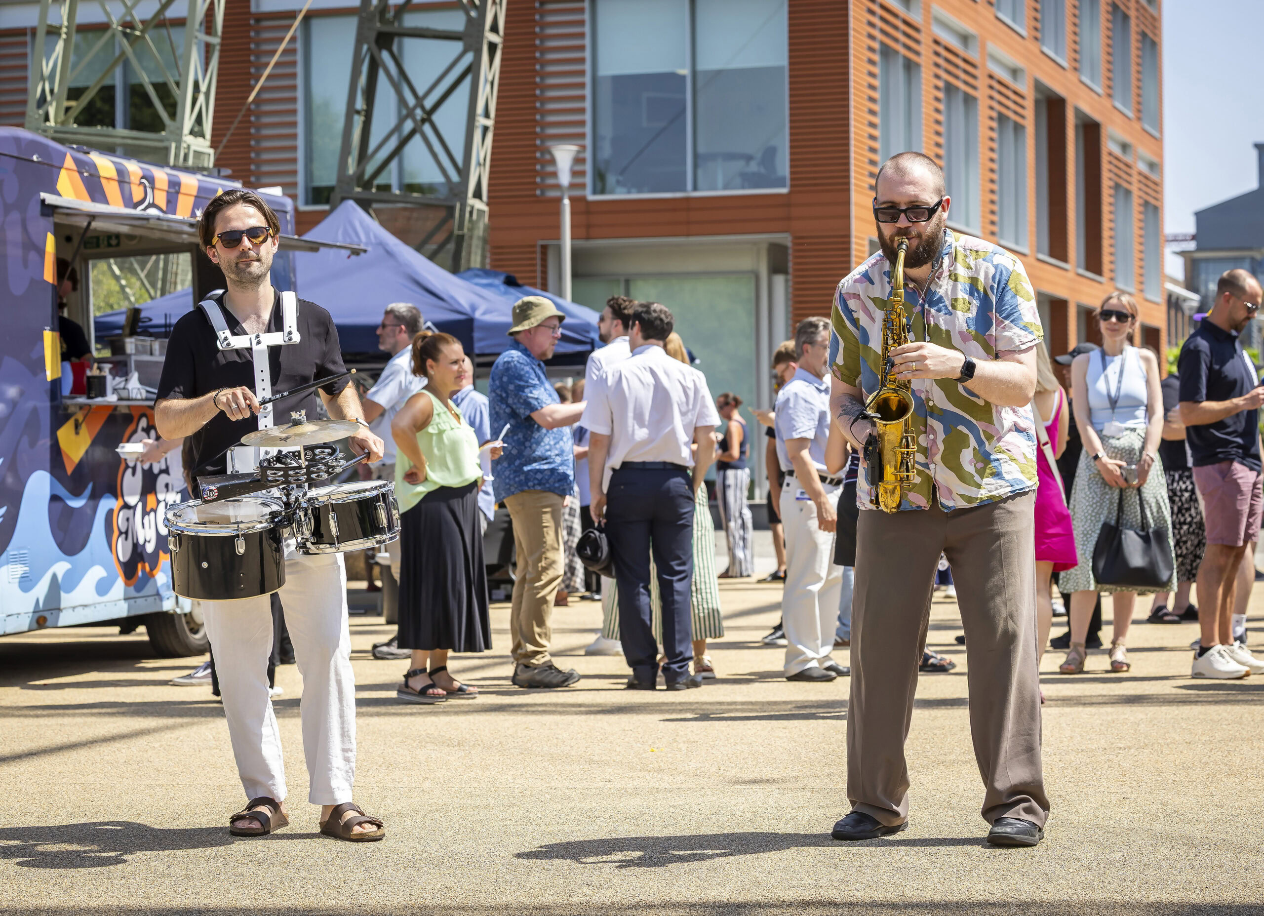 Two musicians, one playing snare drums and one playing saxophone, perform outdoors for a crowd.