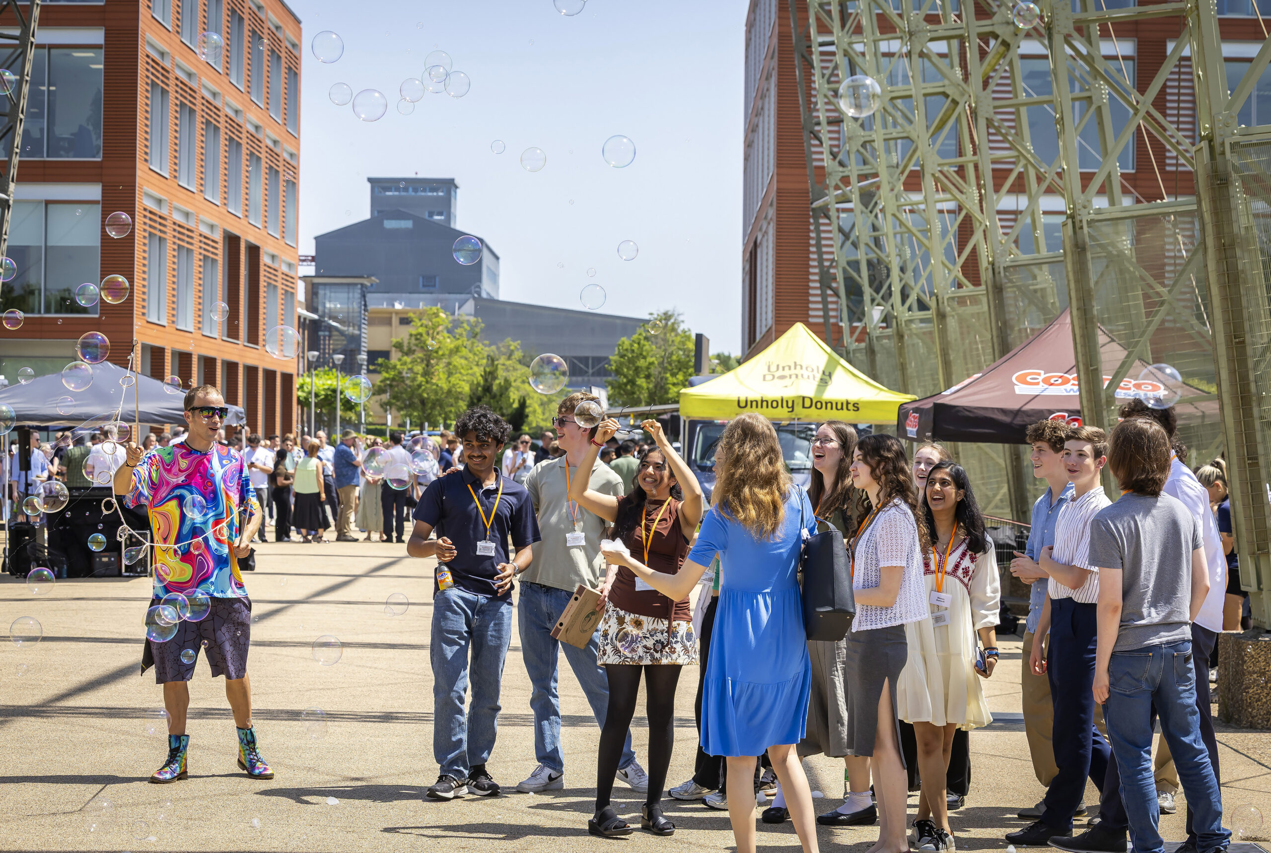 A group of people socialise outdoors near vendor marquees, with bubbles floating in the air.