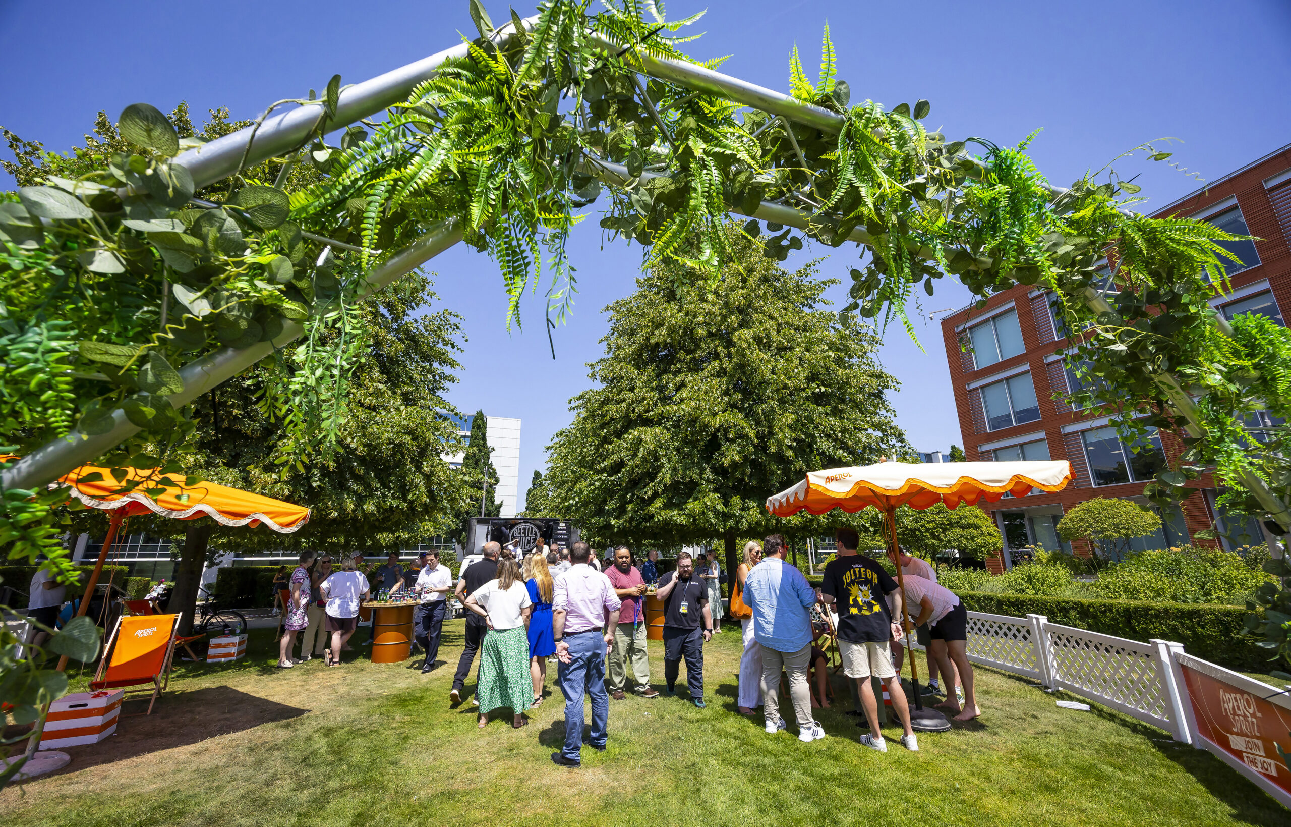 People gather under greenery and orange parasols at an outdoor event on a sunny day.