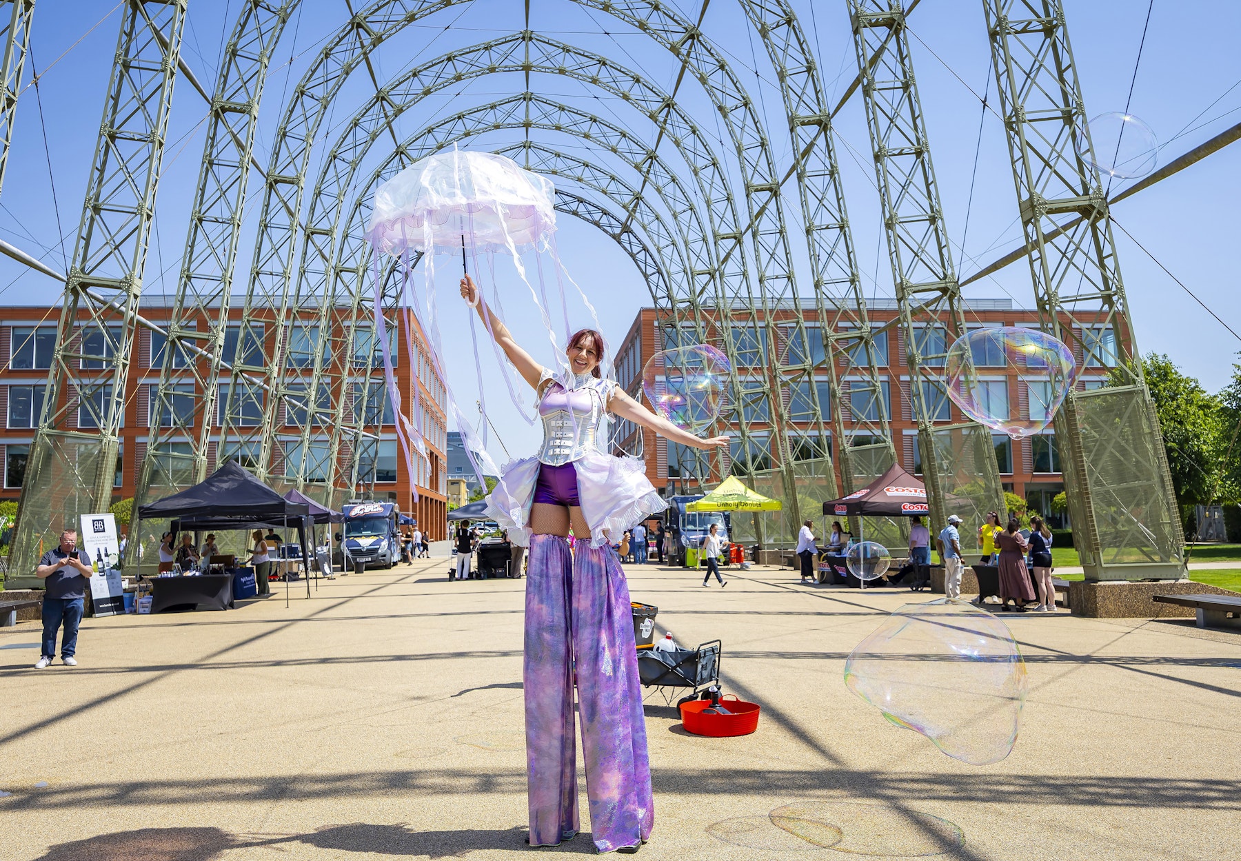 A performer on stilts holds a clear umbrella and large bubbles float in an outdoor event space.